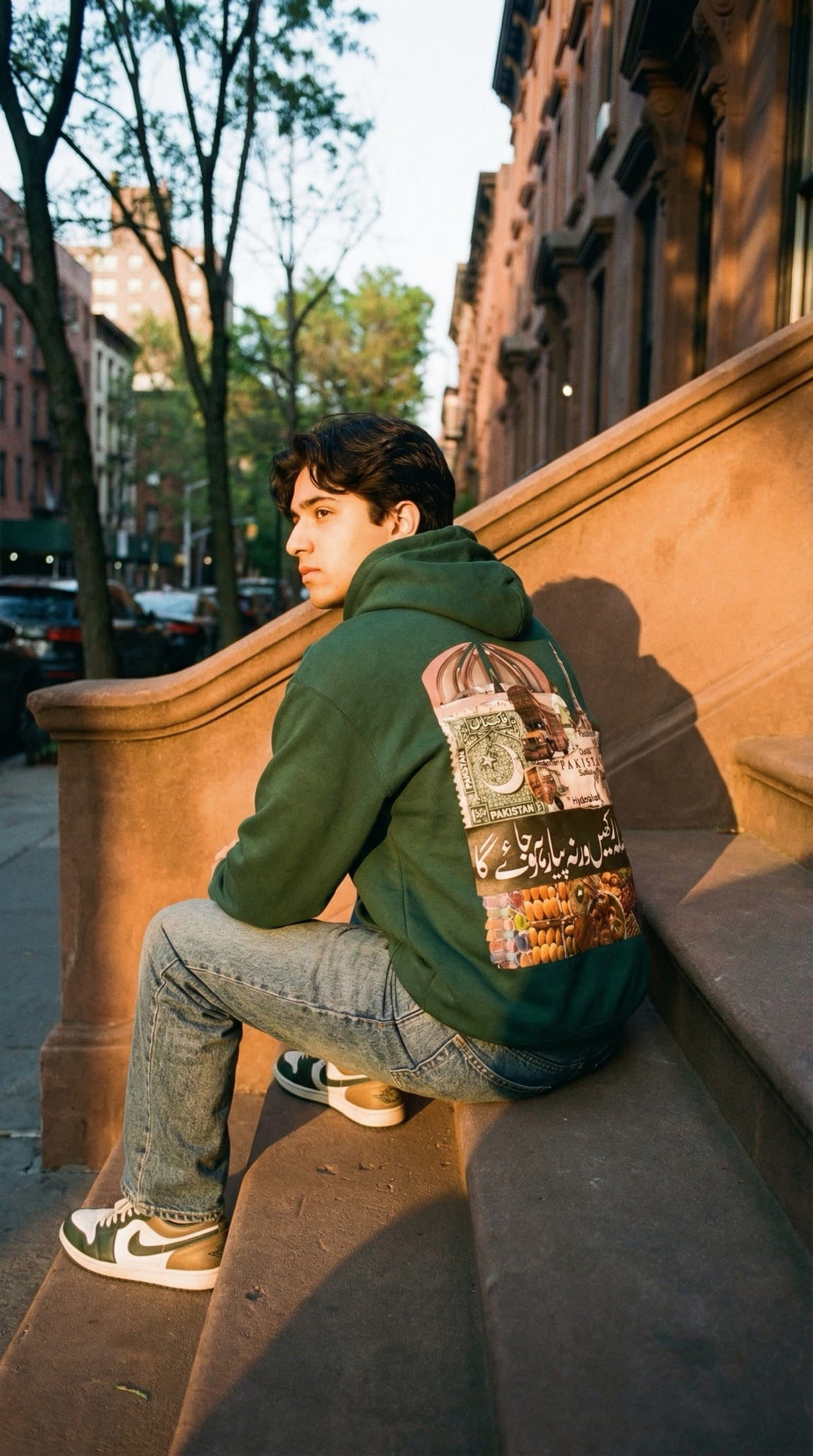 Man in a green Made in Pakistan hoodie sitting on a brownstone stoop