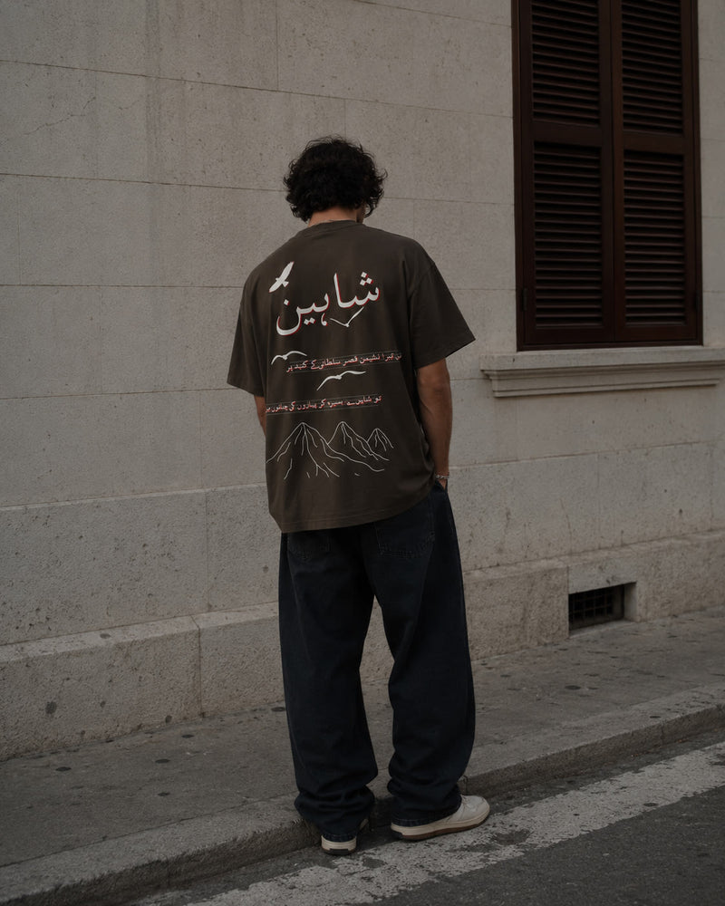 Man in a brown Shaheen tee standing by a stone wall